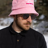 A white man with a beard and brown sunglasses wears a light pink bucket hat with a white illustration of the sun and pink text that says "Unlimbited." He's in a snowy forest.