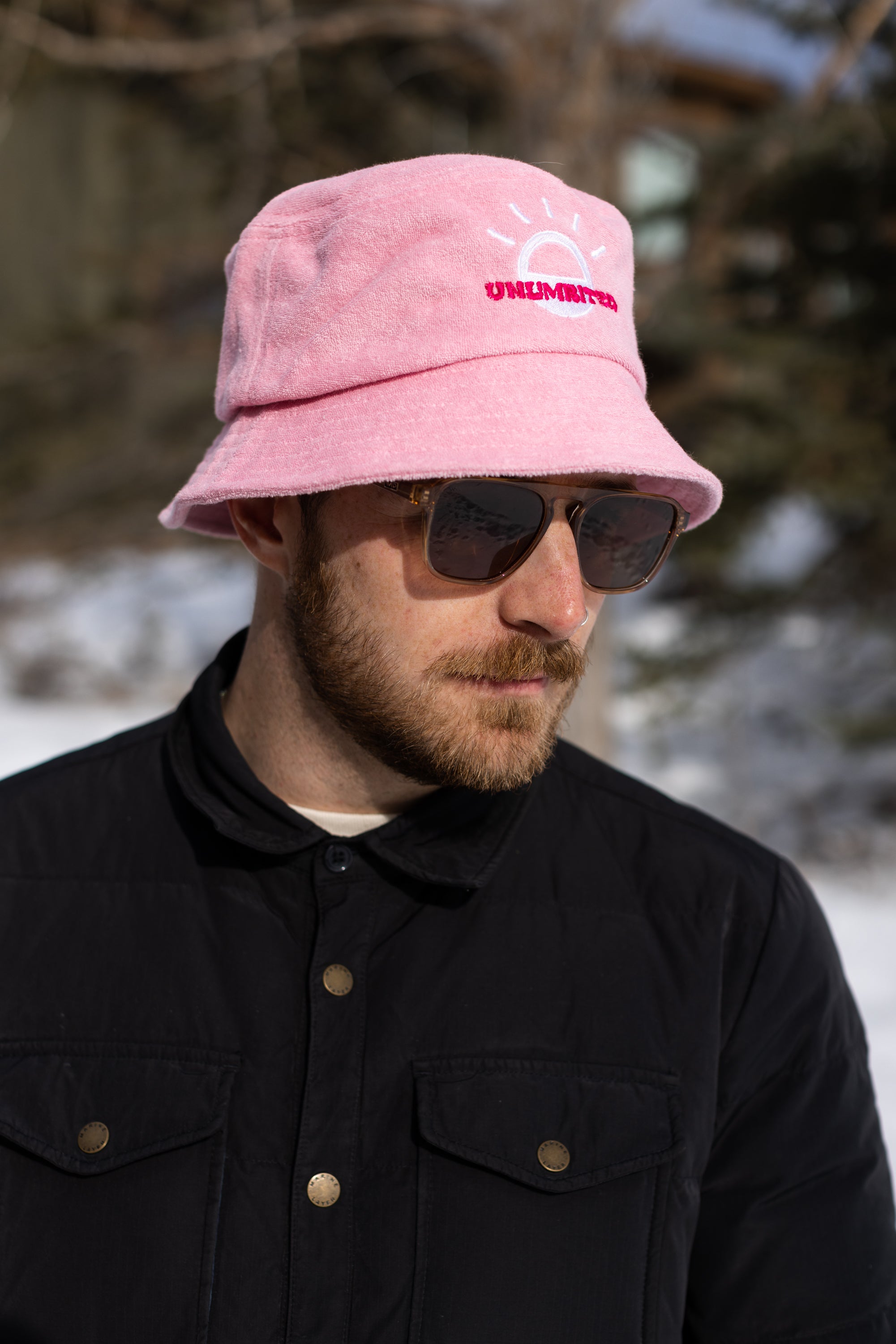 A white man with a beard and brown sunglasses wears a light pink bucket hat with a white illustration of the sun and pink text that says "Unlimbited." He's in a snowy forest.