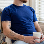 A white man with short brown hair wears a navy t-shirt. He sits on a window seat, holding a mug of coffee.