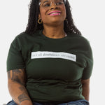 A Black woman (with long black locs, pink lipstick, hoop earrings) wears a forest green short sleeve tee has a beige block printed on the front chest. The words "Not All Disabilities are Visible" are on this block.