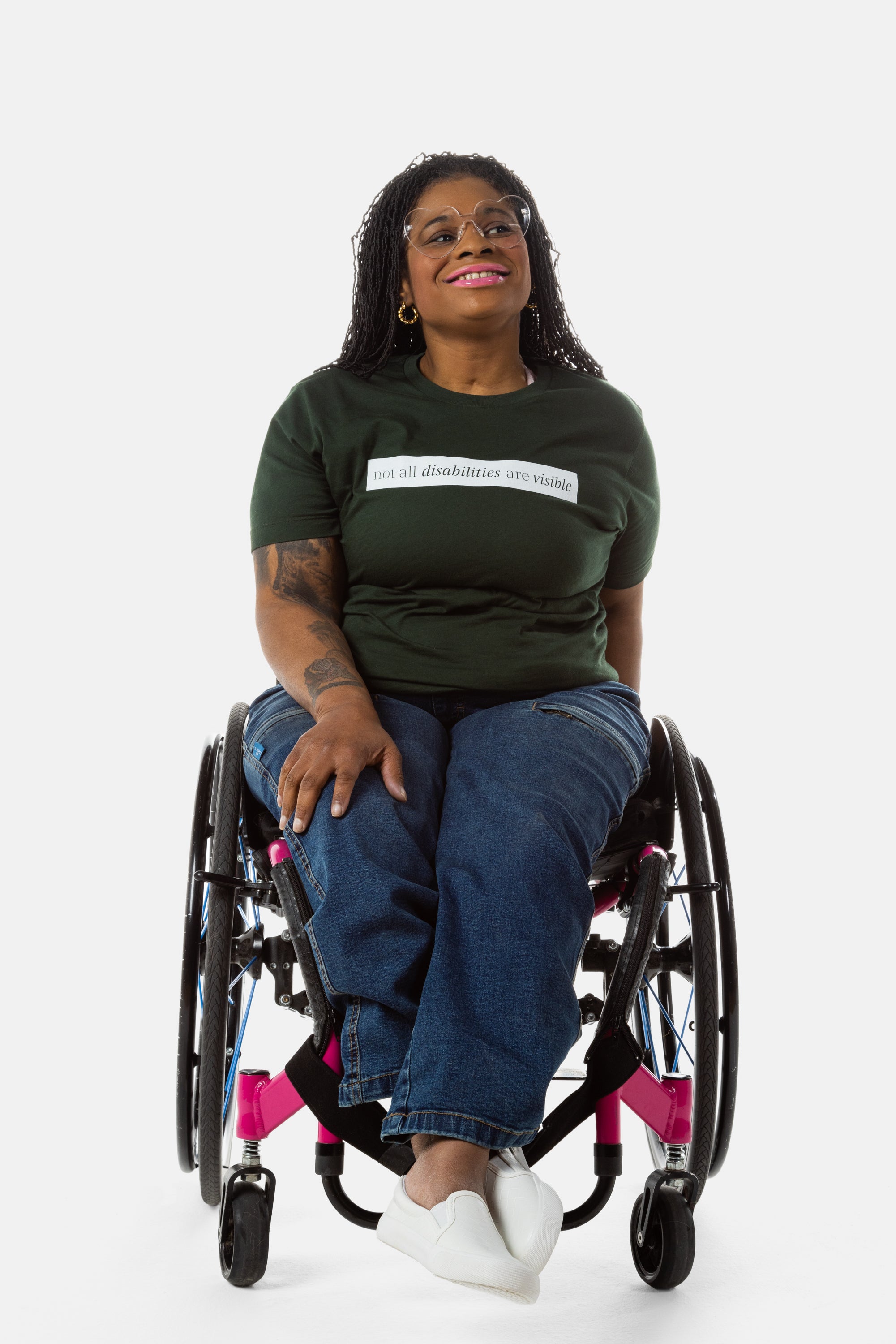 Princess, a Black woman on a pink wheelchair (with long locs, light pink lipstick, clear black sunglasses, and hoops) wears the No Limbits Adaptive Women's Dark Wash Wheelchair Pants.