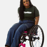 Princess, a Black woman on a pink wheelchair (with long locs, light pink lipstick, clear black sunglasses, and hoops) wears the No Limbits Adaptive Women's Dark Wash Wheelchair Pants.