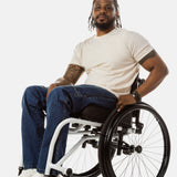 Jessi, a Black man on a white wheelchair (with short locs with a bun on the top of his hair, a goatee, cross earrings, and white sneakers) wears the No Limbits Adaptive Men's Dark Wash Wheelchair Pants.