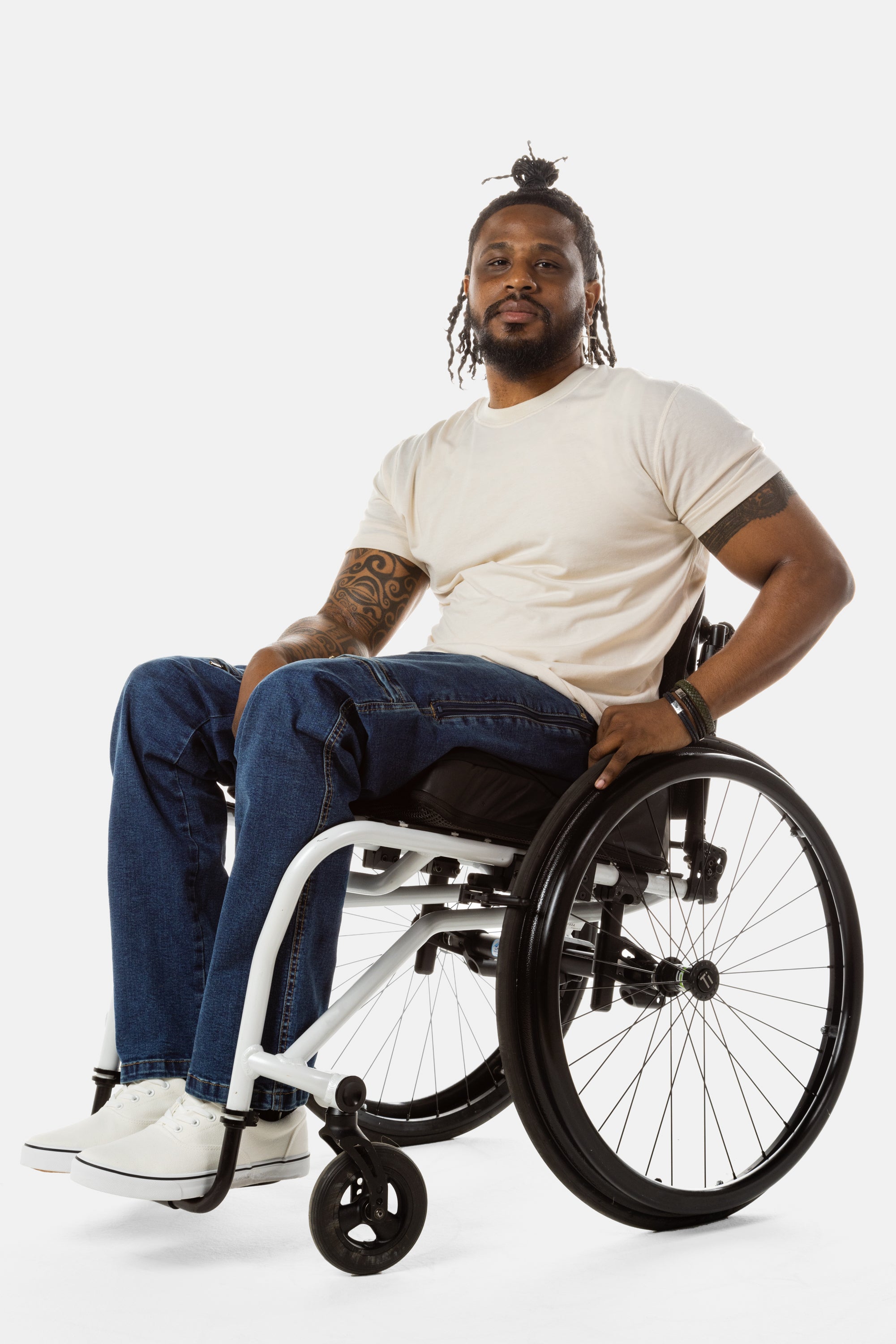 Jessi, a Black man on a white wheelchair (with short locs with a bun on the top of his hair, a goatee, cross earrings, and white sneakers) wears the No Limbits Adaptive Men's Dark Wash Wheelchair Pants.