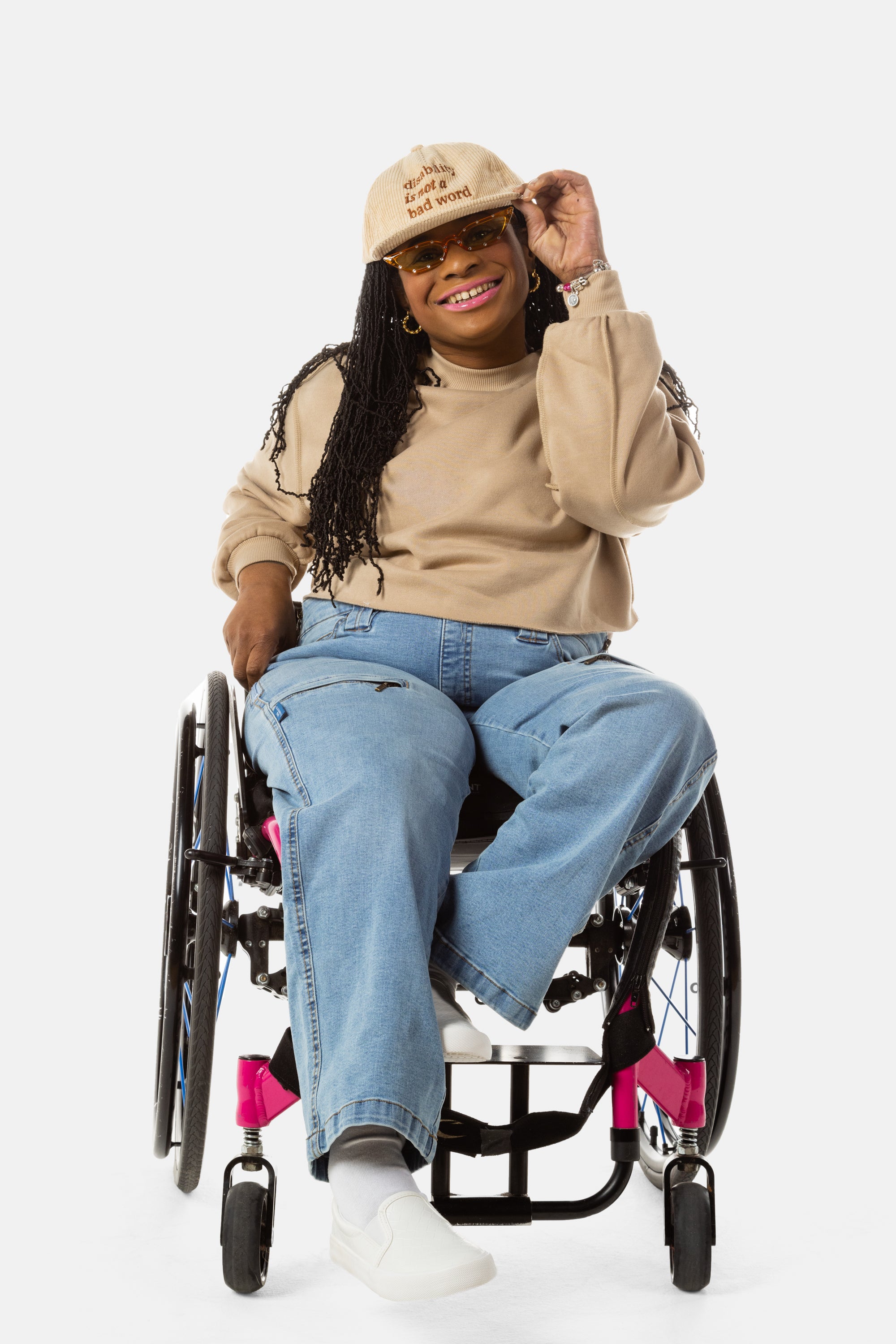 Princess, a Black woman on a pink wheelchair (with long locs, light pink lipstick, clear black sunglasses, and hoops) wears the No Limbits Adaptive Women's Light Wash Wheelchair Pants.