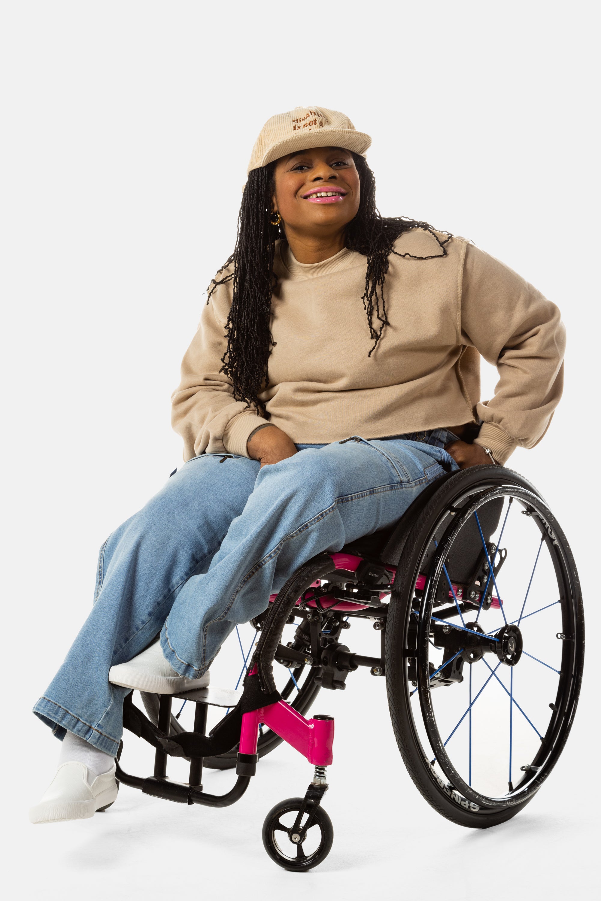 Princess, a Black woman on a pink wheelchair (with long locs, light pink lipstick, clear black sunglasses, and hoops) wears the No Limbits Adaptive Women's Light Wash Wheelchair Pants.