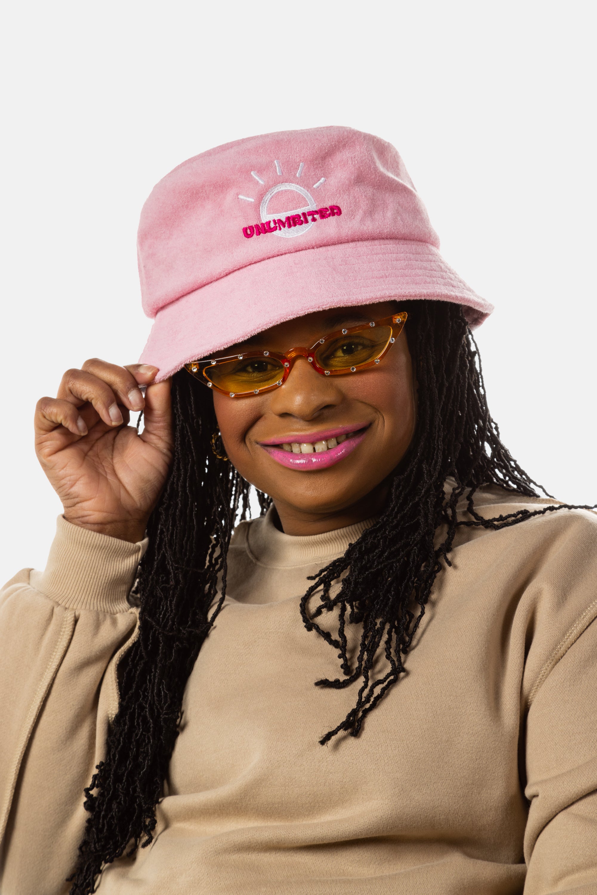 A black woman with sunglasses, pink lipstick, and long black locs wears a light pink bucket hat with a white illustration of the sun and pink text that says "Unlimbited"