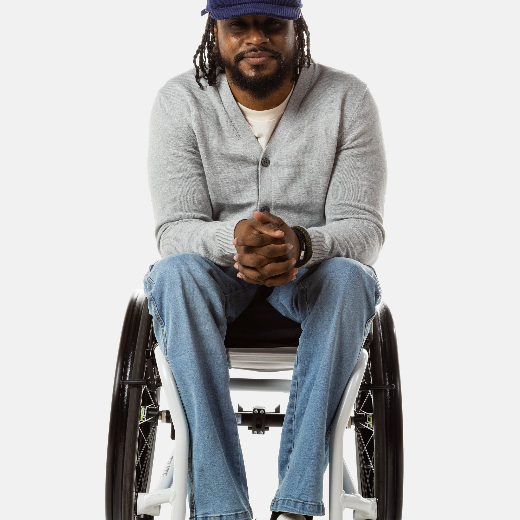 Jessi, a Black man on a white wheelchair (with short locs with a bun on the top of his hair, a goatee, cross earrings, and white sneakers) wears the No Limbits Adaptive Men's Light Wash Wheelchair Pants.