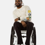 Jessi, a Black man on a white wheelchair (with short locs with a bun on the top of his hair, a goatee, cross earrings, and white sneakers) wears the No Limbits Adaptive Men's Black Wheelchair Pants.