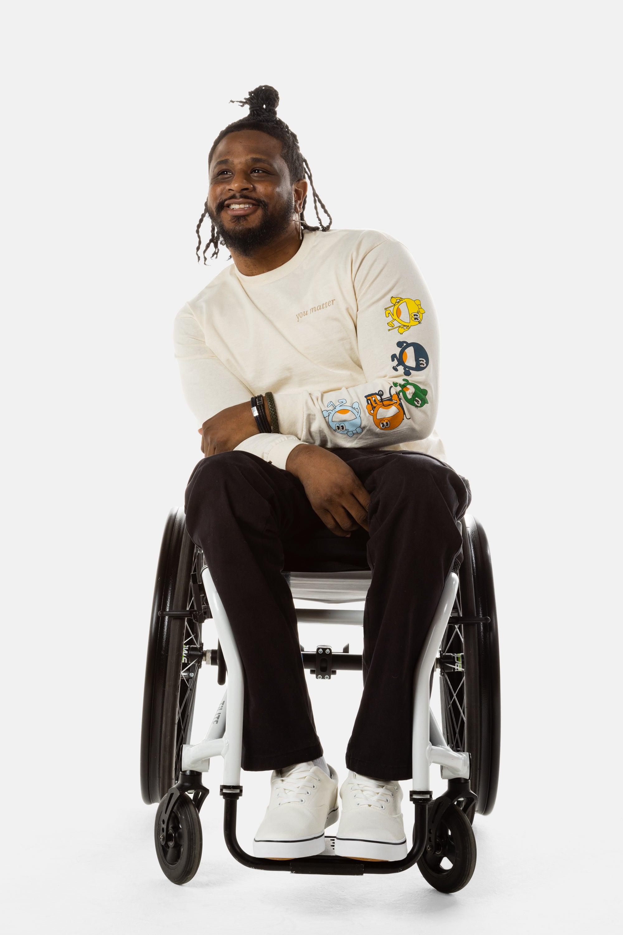 Jessi, a Black man on a white wheelchair (with short locs with a bun on the top of his hair, a goatee, cross earrings, and white sneakers) wears the No Limbits Adaptive Men's Black Wheelchair Pants.