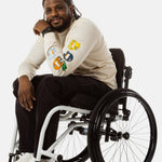 Jessi, a Black man on a white wheelchair (with short locs with a bun on the top of his hair, a goatee, cross earrings, and white sneakers) wears the No Limbits Adaptive Men's Black Wheelchair Pants.