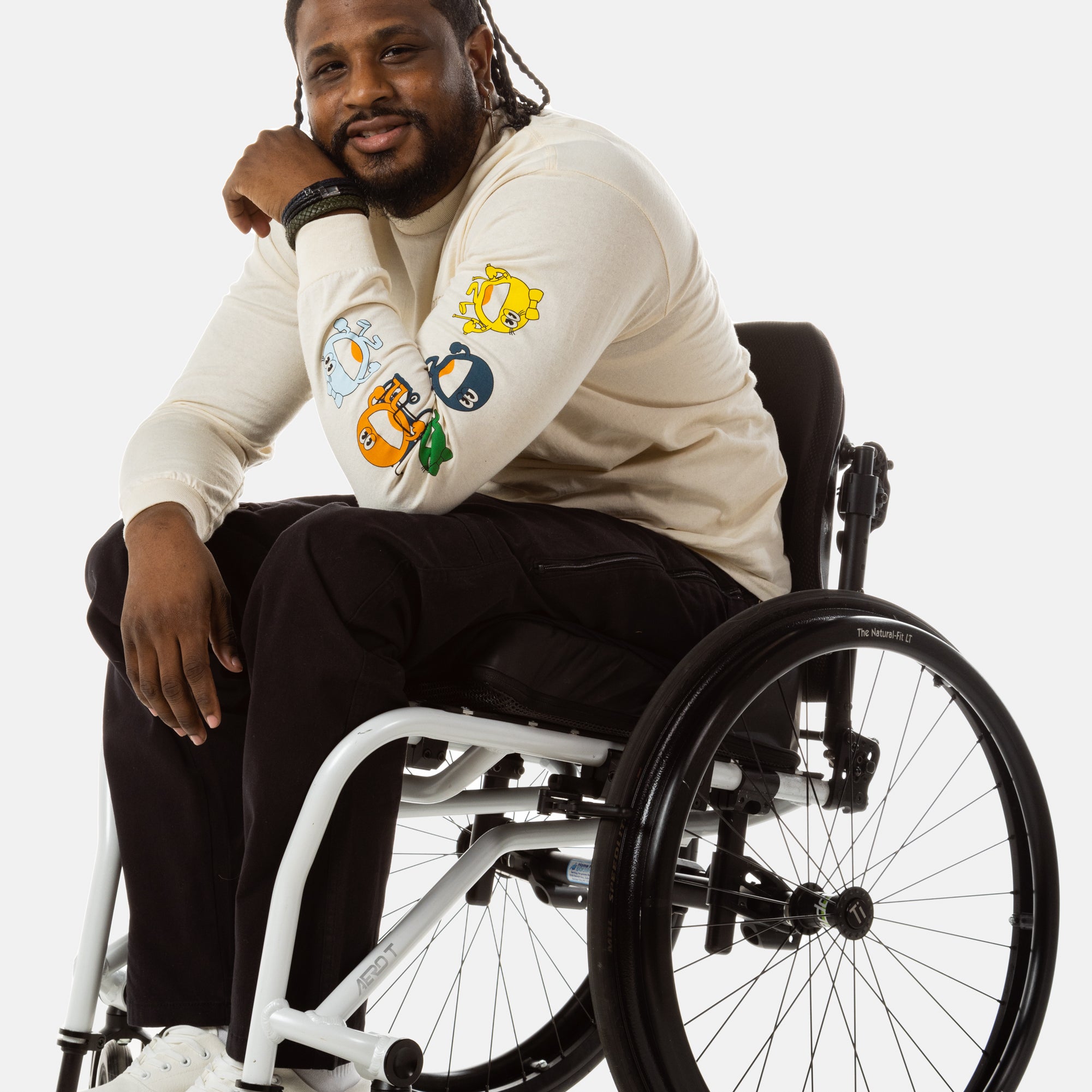 Jessi, a Black man on a white wheelchair (with short locs with a bun on the top of his hair, a goatee, cross earrings, and white sneakers) wears the No Limbits Adaptive Men's Black Wheelchair Pants.