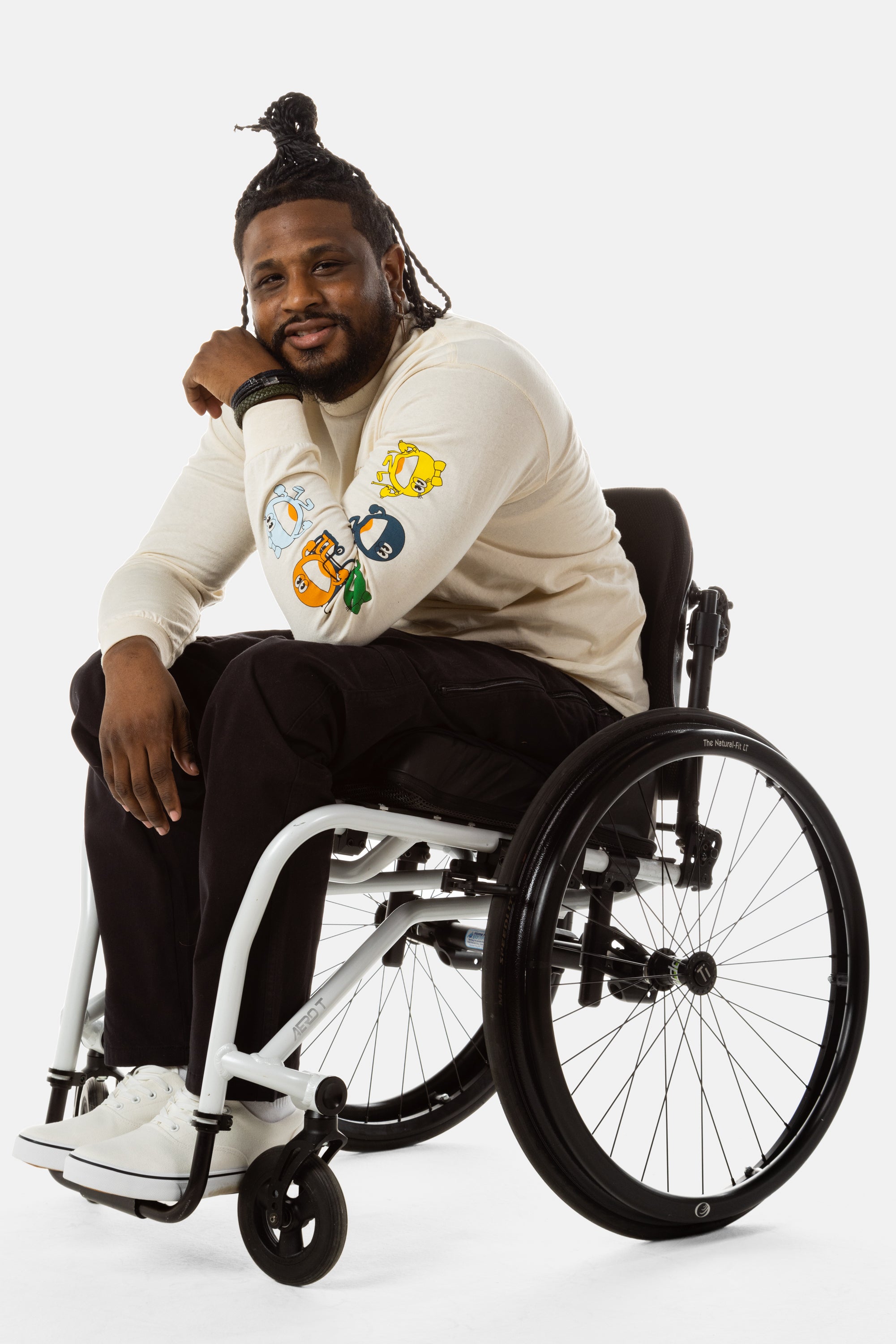Jessi, a Black man on a white wheelchair (with short locs with a bun on the top of his hair, a goatee, cross earrings, and white sneakers) wears the No Limbits Adaptive Men's Black Wheelchair Pants.