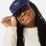 A black woman with sunglasses, pink lipstick, and long black locs wears a dark blue baseball cap that has a illustration of the moon and the stars that says in white font "Sky's the Limbit."