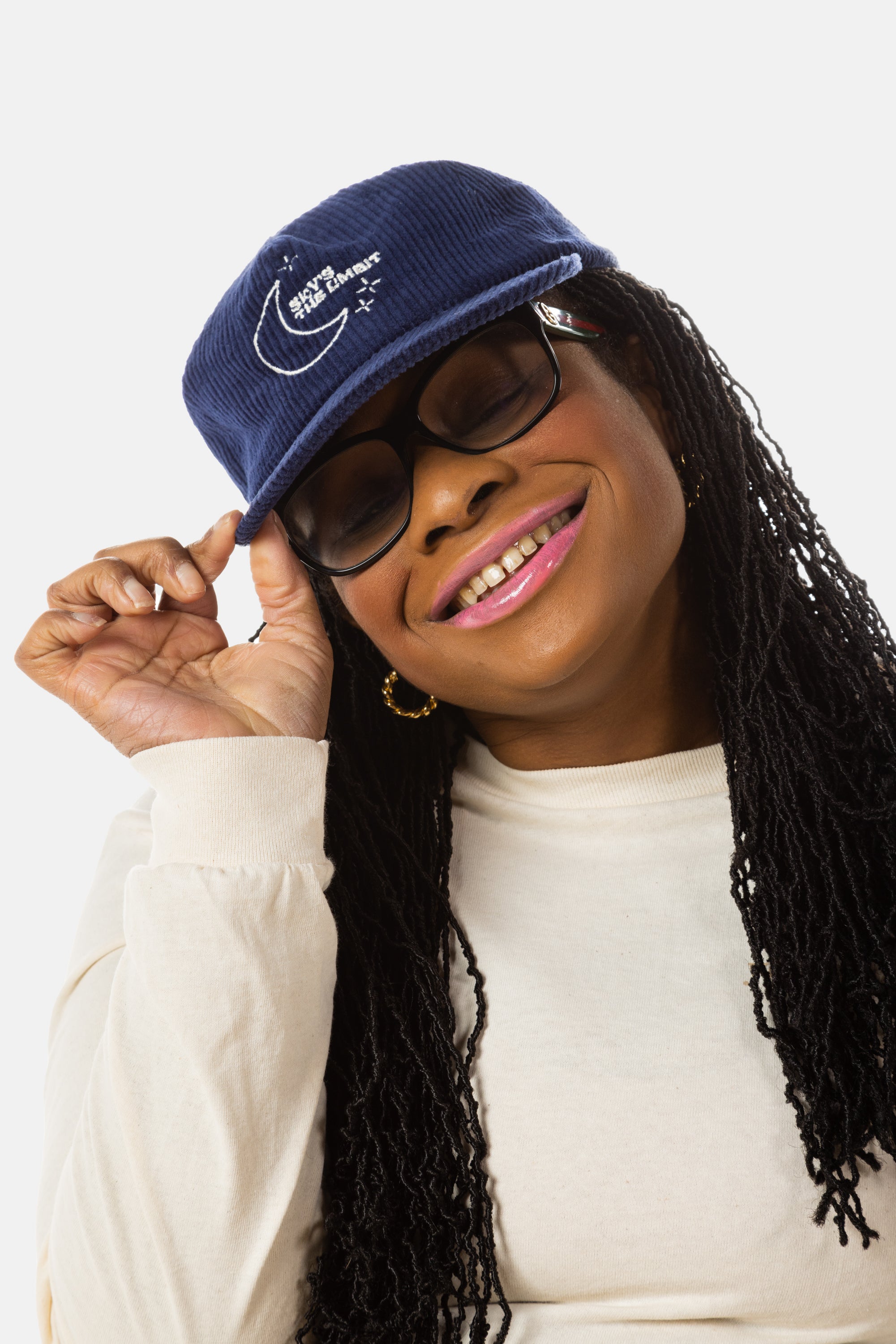 A black woman with sunglasses, pink lipstick, and long black locs wears a dark blue baseball cap that has a illustration of the moon and the stars that says in white font "Sky's the Limbit."