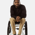 Jessi, a Black man on a white wheelchair (with short locs with a bun on the top of his hair, a goatee, cross earrings, and white sneakers) wears the No Limbits Adaptive Men's Khaki Wheelchair Pants.