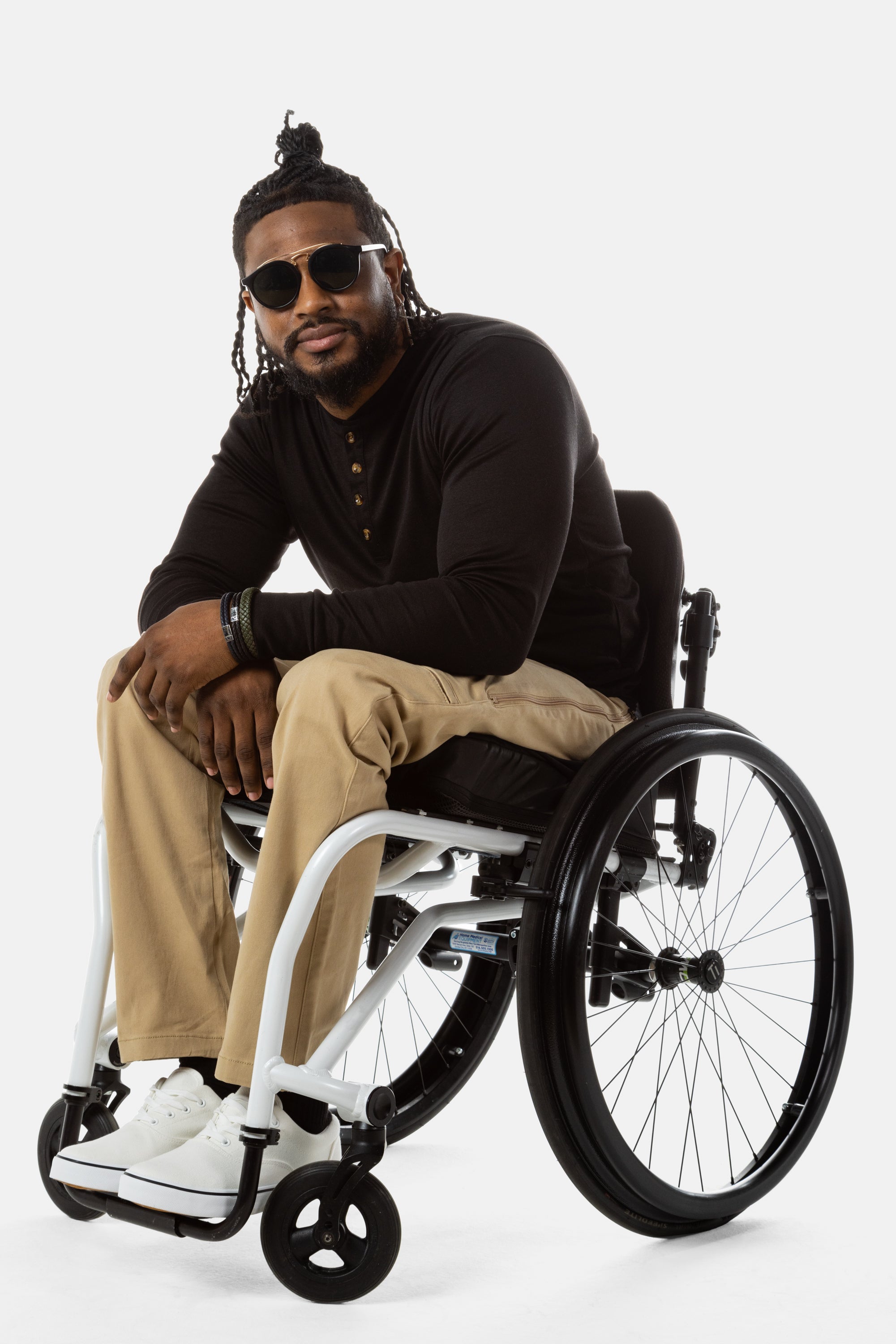 Jessi, a Black man on a white wheelchair (with short locs with a bun on the top of his hair, a goatee, cross earrings, and white sneakers) wears the No Limbits Adaptive Men's Khaki Wheelchair Pants. He is wearing black sunglasses.