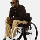 Jessi, a Black man on a white wheelchair (with short locs with a bun on the top of his hair, a goatee, cross earrings, and white sneakers) wears the No Limbits Adaptive Men's Khaki Wheelchair Pants. He is wearing black sunglasses.