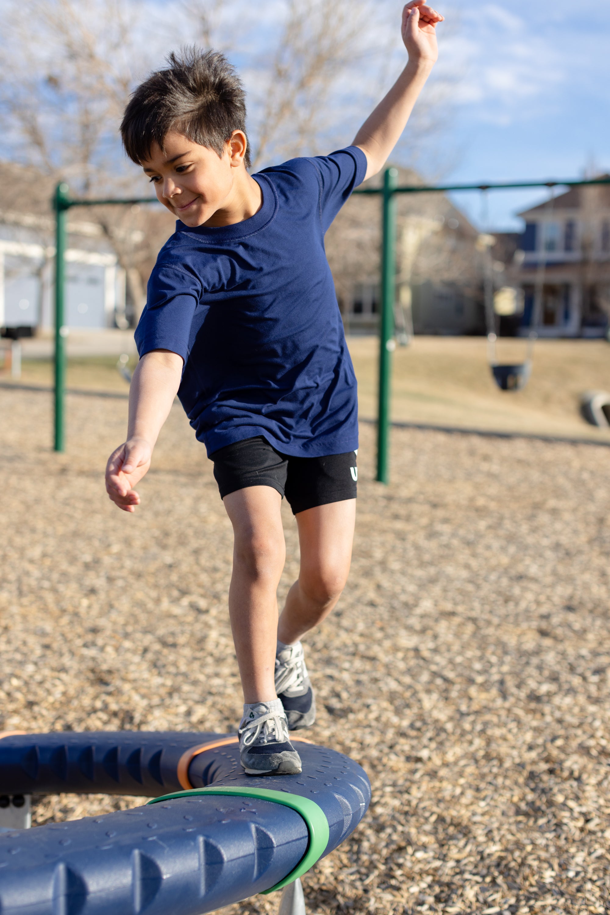 A little boy playing in the park, he is wearing a navy basic crewneck t-shirt and black shorts. You can see swing sets blurred out in the background of the photo.