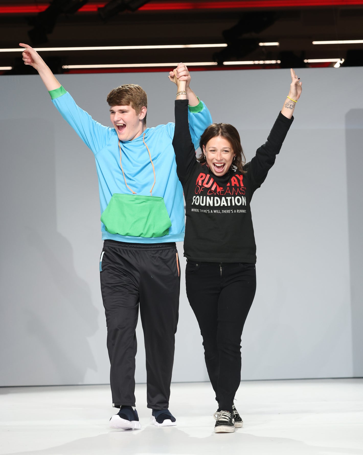 A photo of a male model wearing black sweatpants and a light blue and green sweatshirt. He is being assisted with a female who is wearing all black, and they are walking on an all white runway.