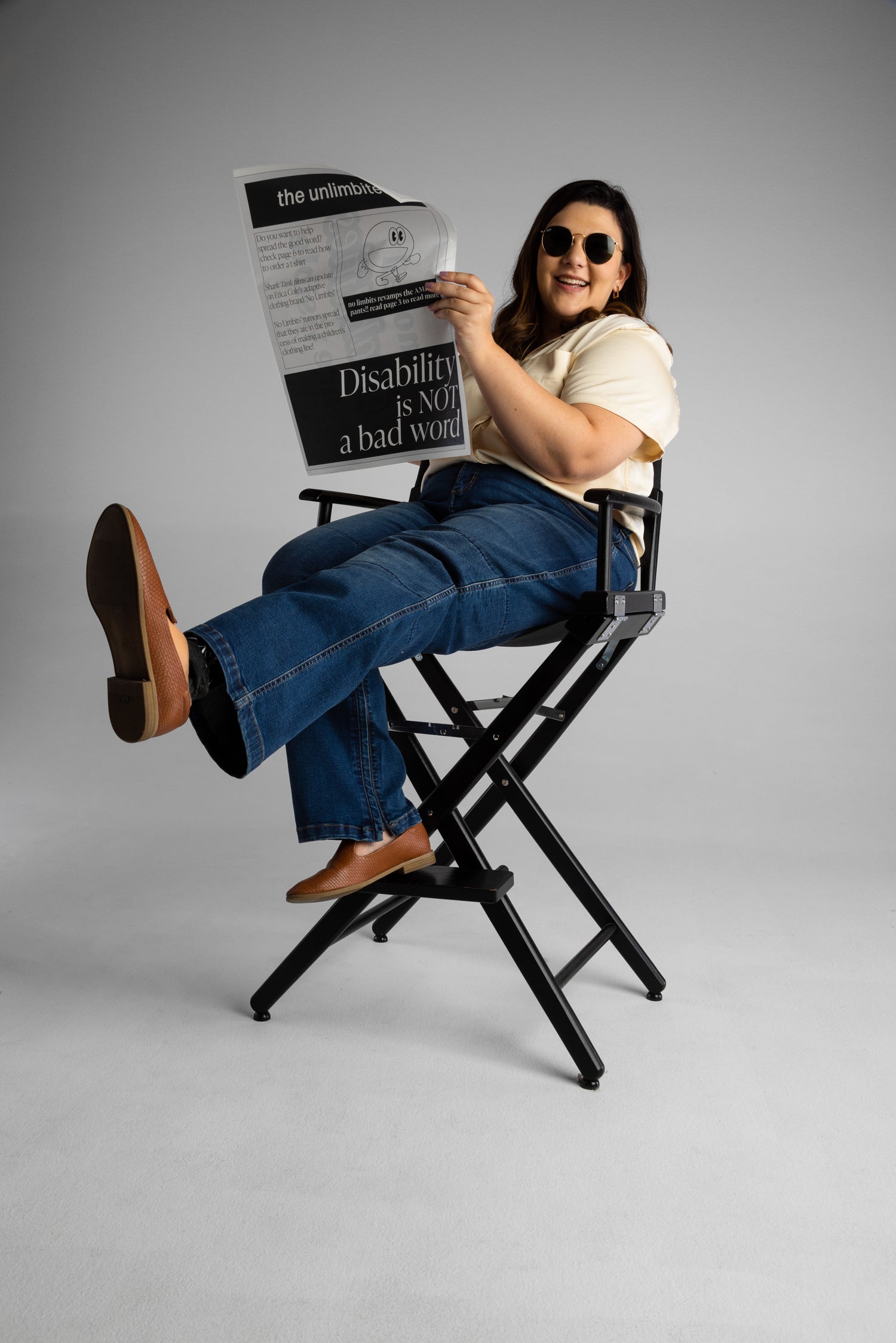 Erica is posing while sitting on a black directors chair. She has one leg kicked out so you can see her brown boots. She is wearing dark denim jeans and a tan blouse, and is holding a newspaper that reads 'Disability is not a bad word'