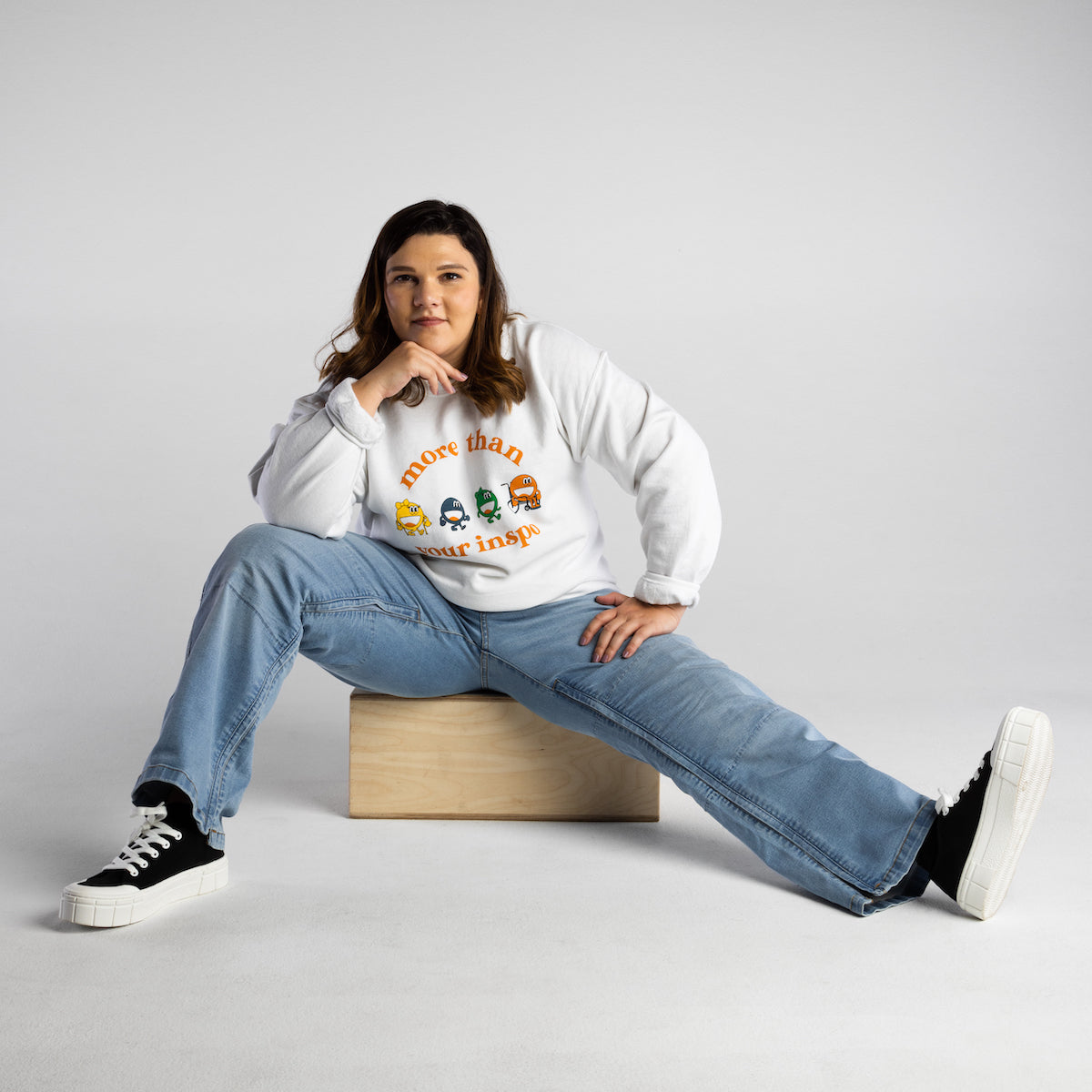 Erica is sitting on top of a tan box and is wearing light wash denim and a white long sleeve shirt with a graphic on it. She is also wearing black and white shoes.