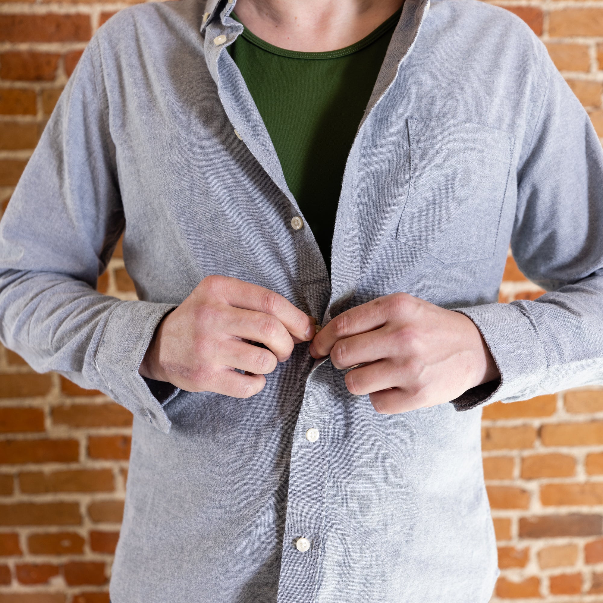 A white man buttons a light denim jacket over his olive-green tank top and denim jeans. He's by a brick background.