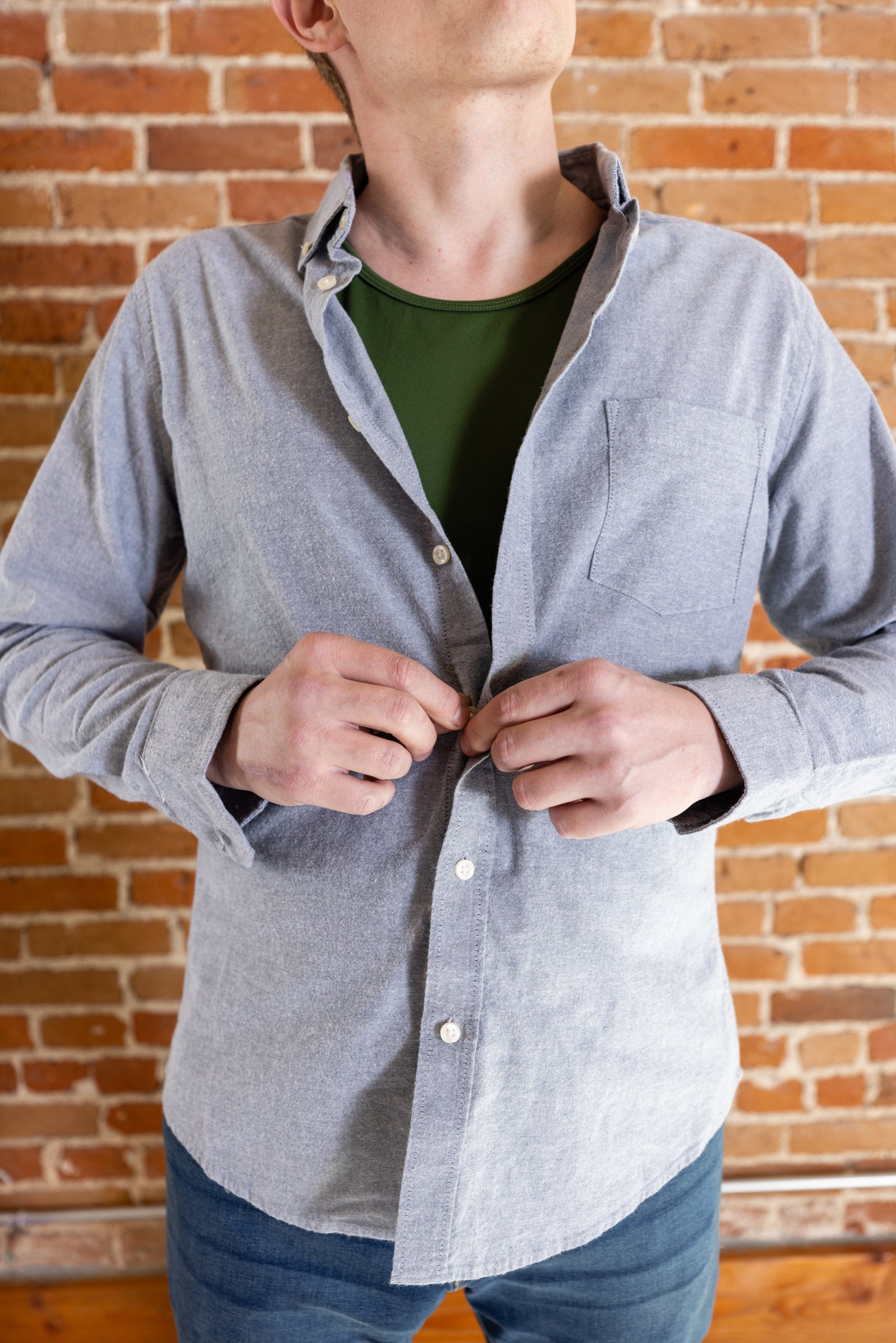 A white man buttons a light denim jacket over his olive-green tank top and denim jeans. He's by a brick background.