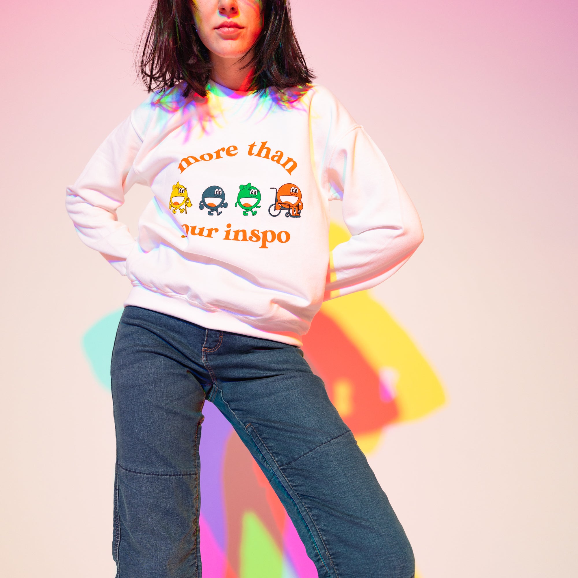 Sophia (A Latina woman with long black hair) poses in front of a white wall. There are outlines behind her that are light blue, purple, pink, and yellow. She is wearing a white sweater that says “not your inspo with the four No Limbits crew members (Colorful circles with forearm crutches, a prosthetic leg, and a wheelchair), and denim jeans.