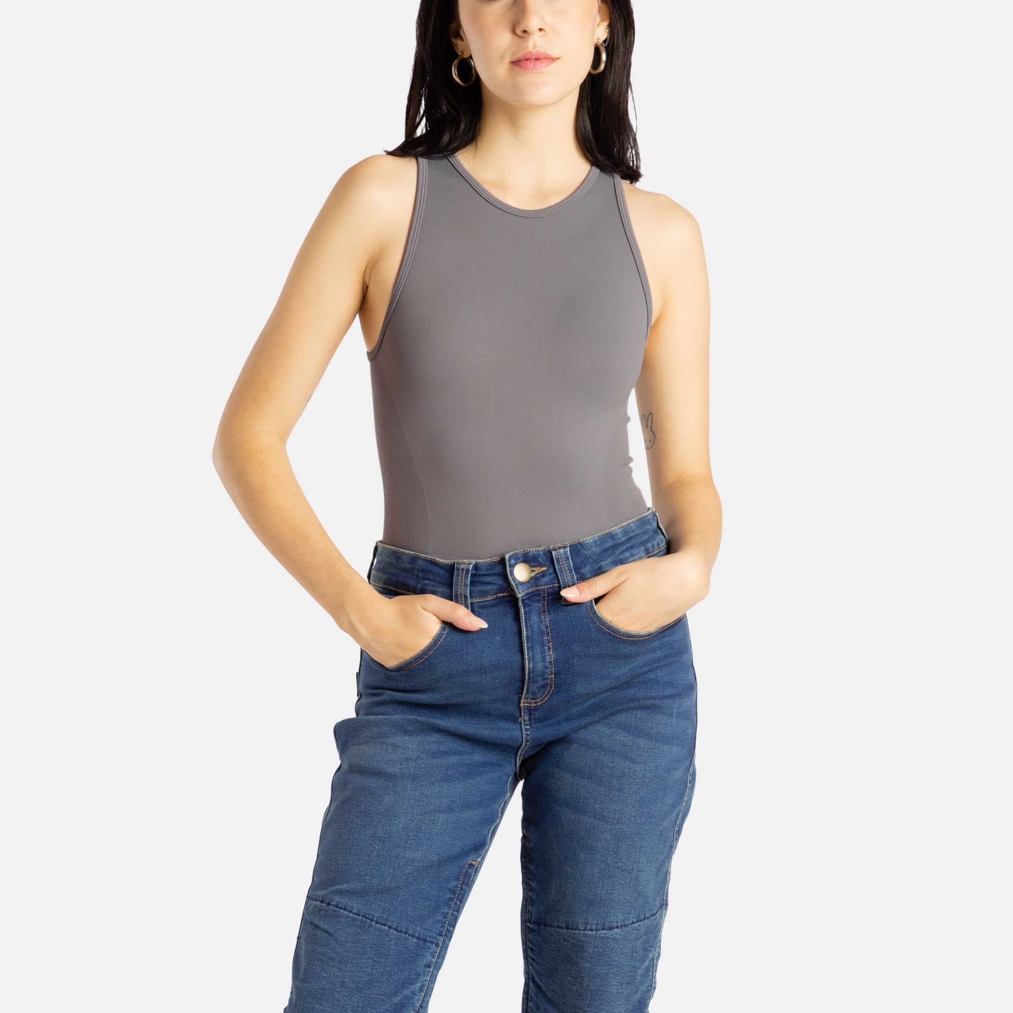 A white woman with long black hair and hoop earrings wears a charcoal tank top and denim jeans.