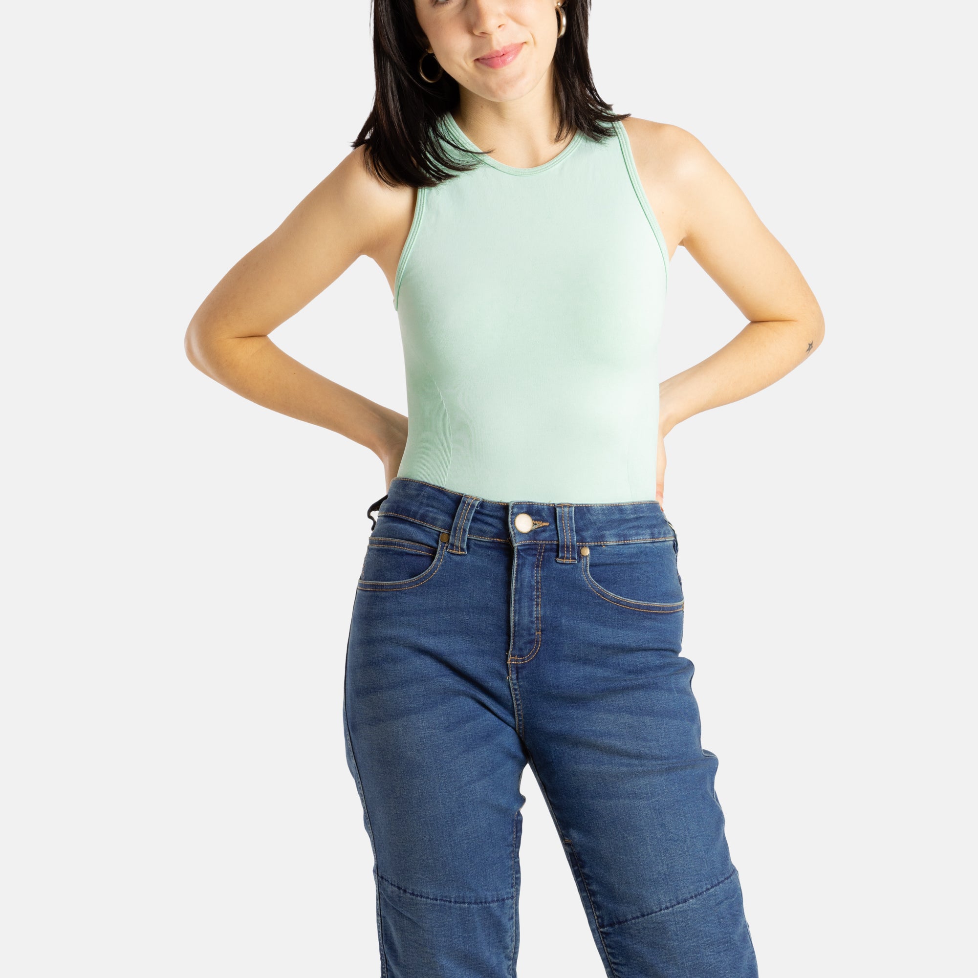 A white woman with long black hair and hoop earrings wears a sea form (Very light green) tank top and denim jeans.