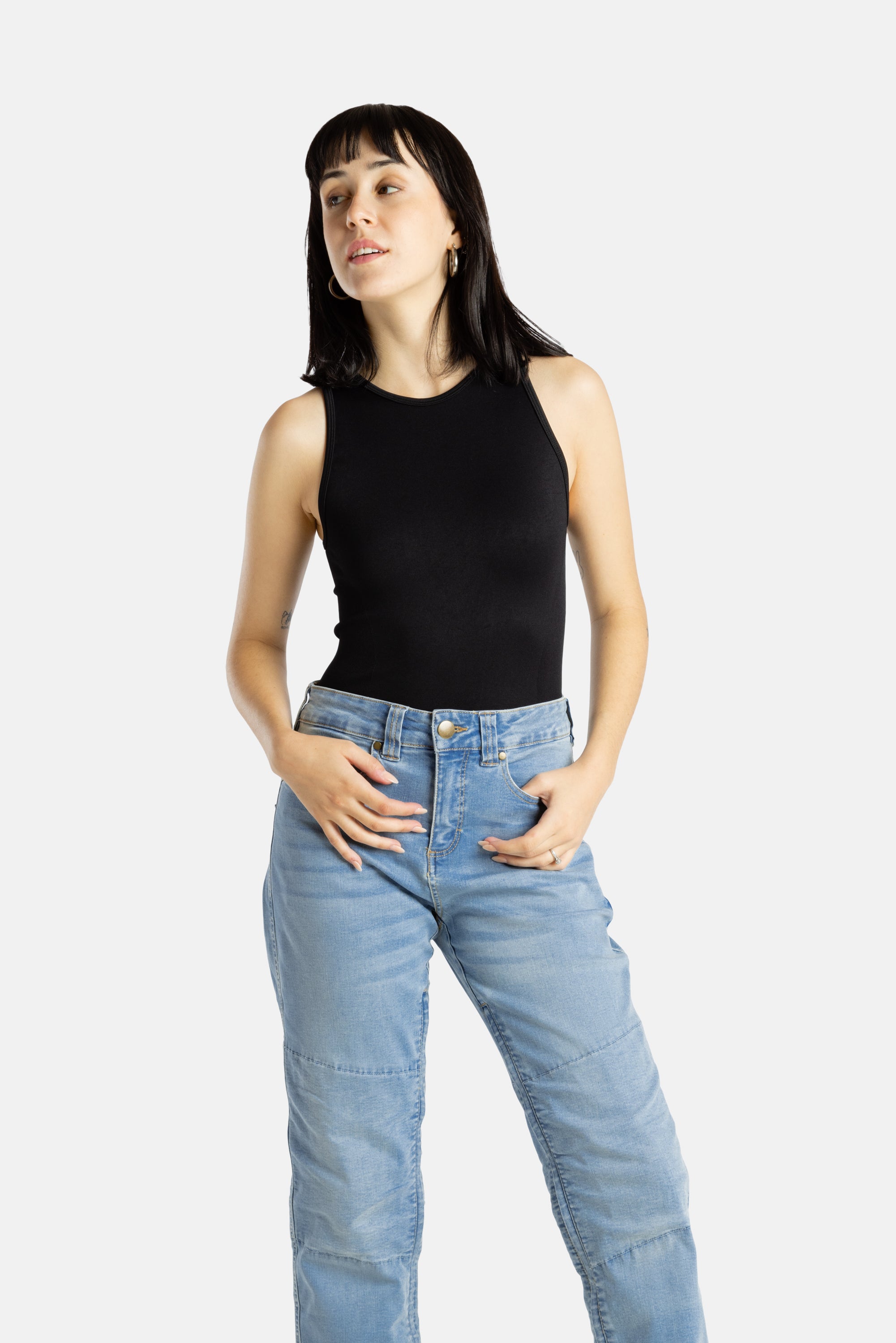 A white woman with long black hair and hoop earrings wears a black tank top and denim jeans.