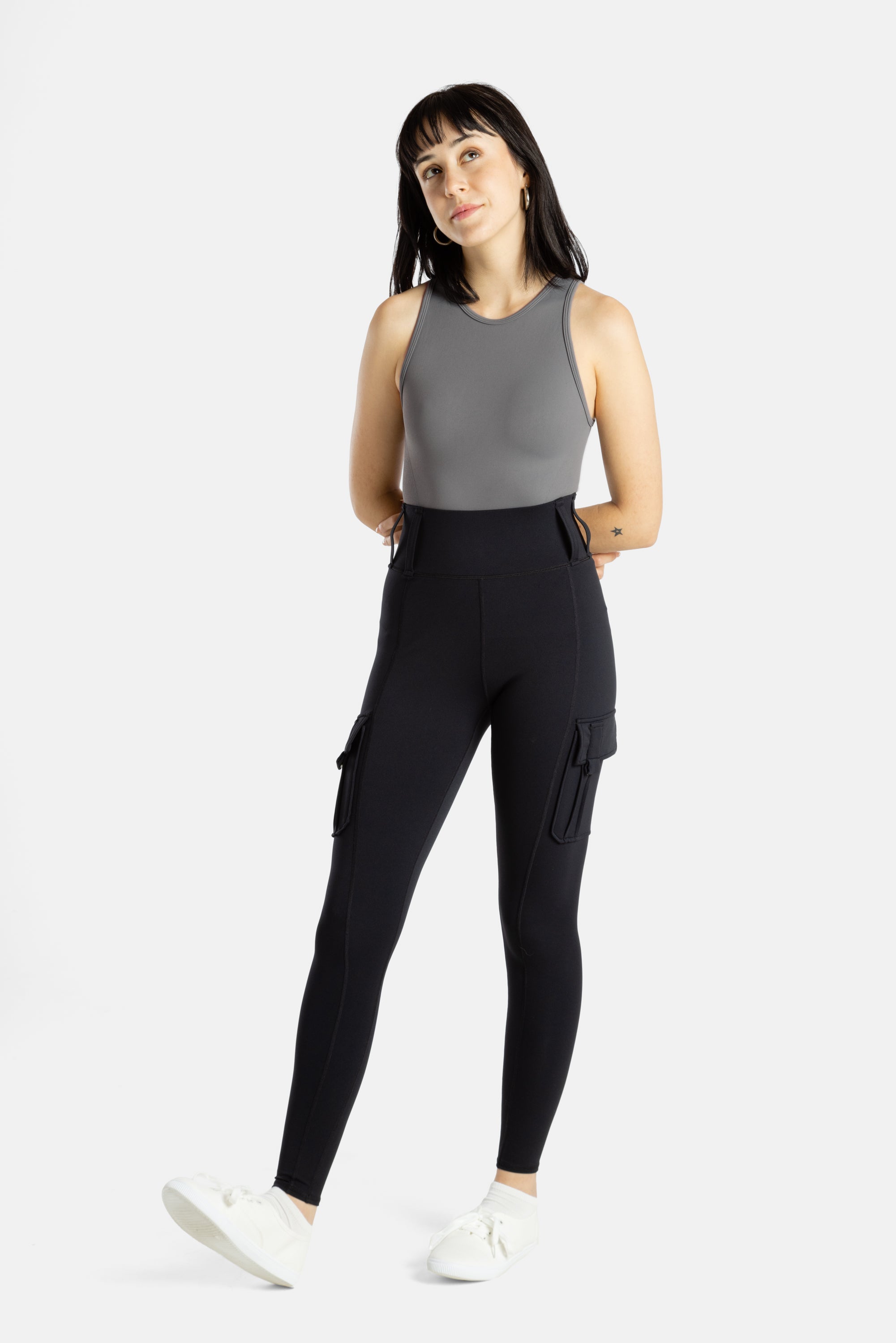 A white woman with long black hair and hoop earrings wears a charcoal tank top and black leggings. Her arms are behind her back and she raises her right ankle, showing her white sneakers.