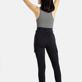 The back of an white woman (with long black hair and hoop earrings) who is wearing a charcoal tank top and black leggings. Her arms are on her head.