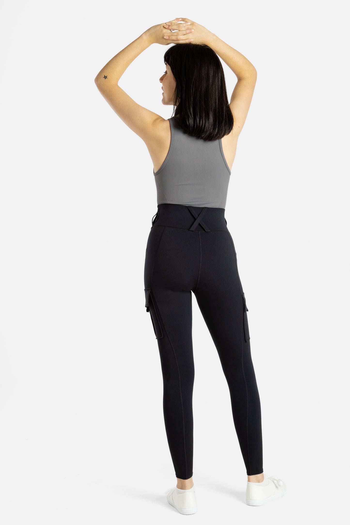 The back of an white woman (with long black hair and hoop earrings) who is wearing a charcoal tank top and black leggings. Her arms are on her head.