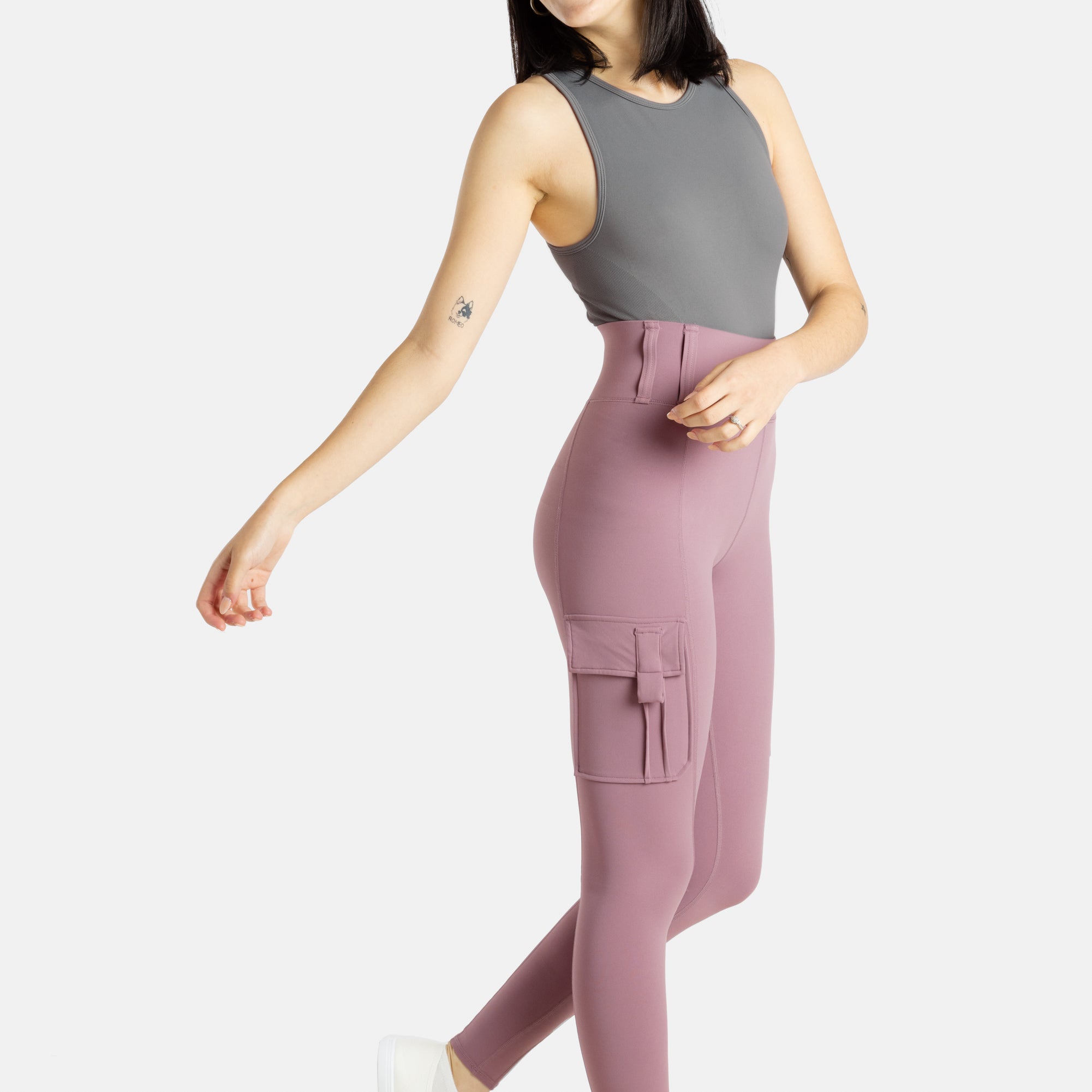 A white woman with long black hair and hoop earrings wears a charcoal tank top and mauve (Pinkish lavender) leggings.