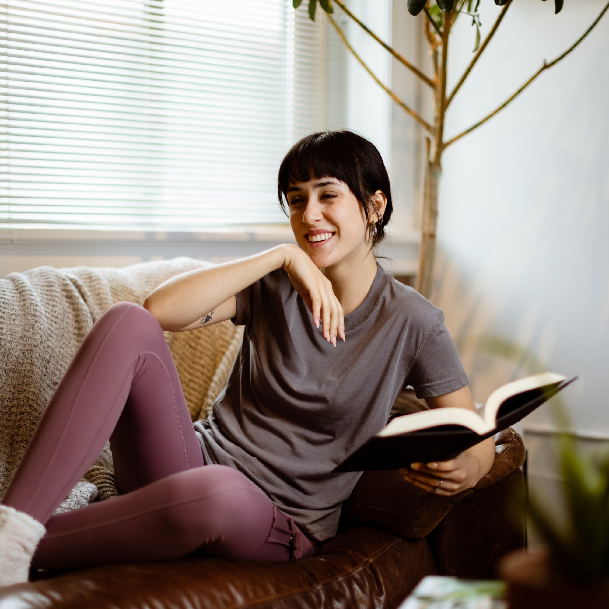 Sophia (A woman with long black hair) wears the No Limbits Adaptive Women's Charcoal Sensory Tee. She lays on a couch, holding a book.