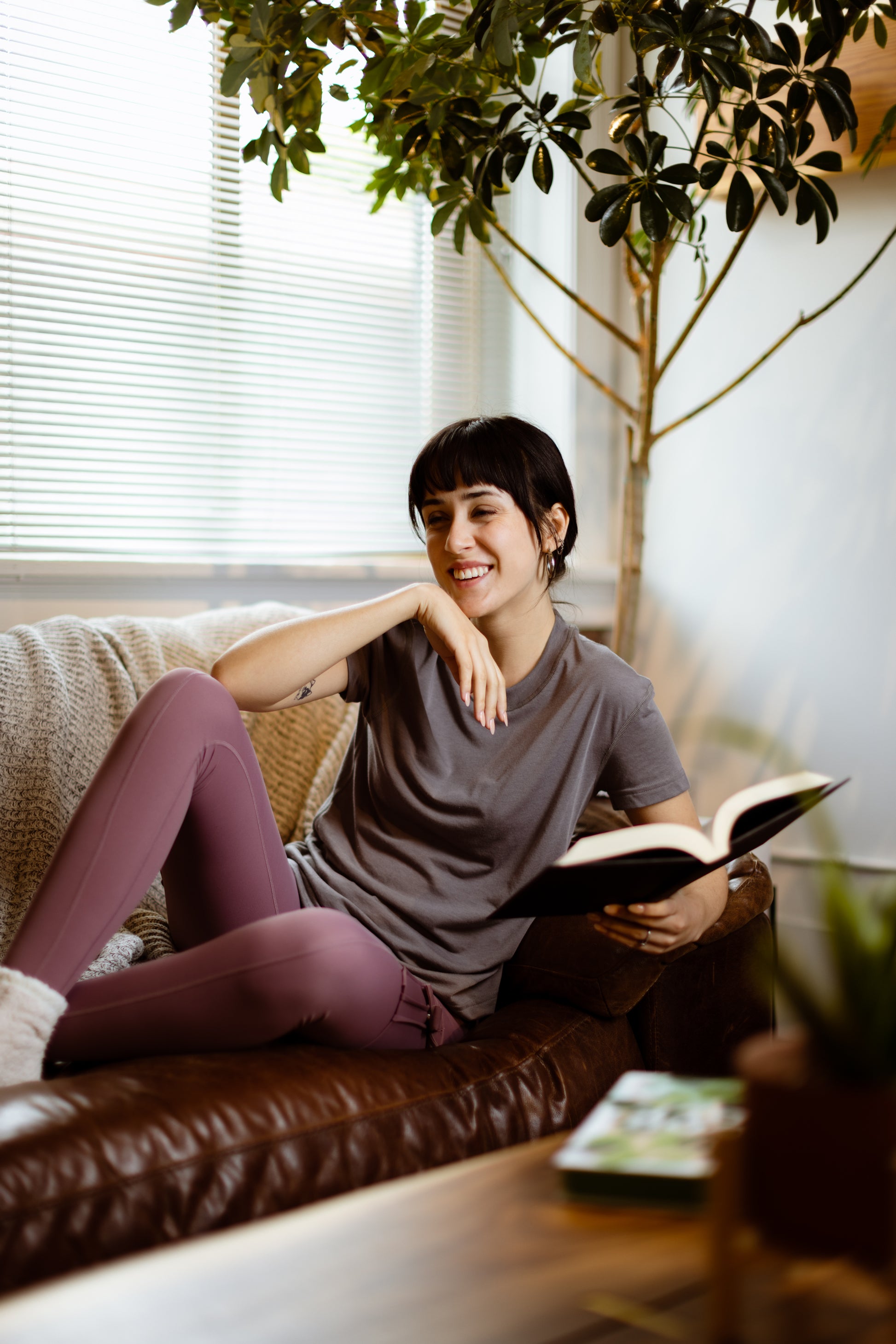 Sophia (A woman with long black hair) wears the No Limbits Adaptive Women's Charcoal Sensory Tee. She lays on a couch, holding a book.
