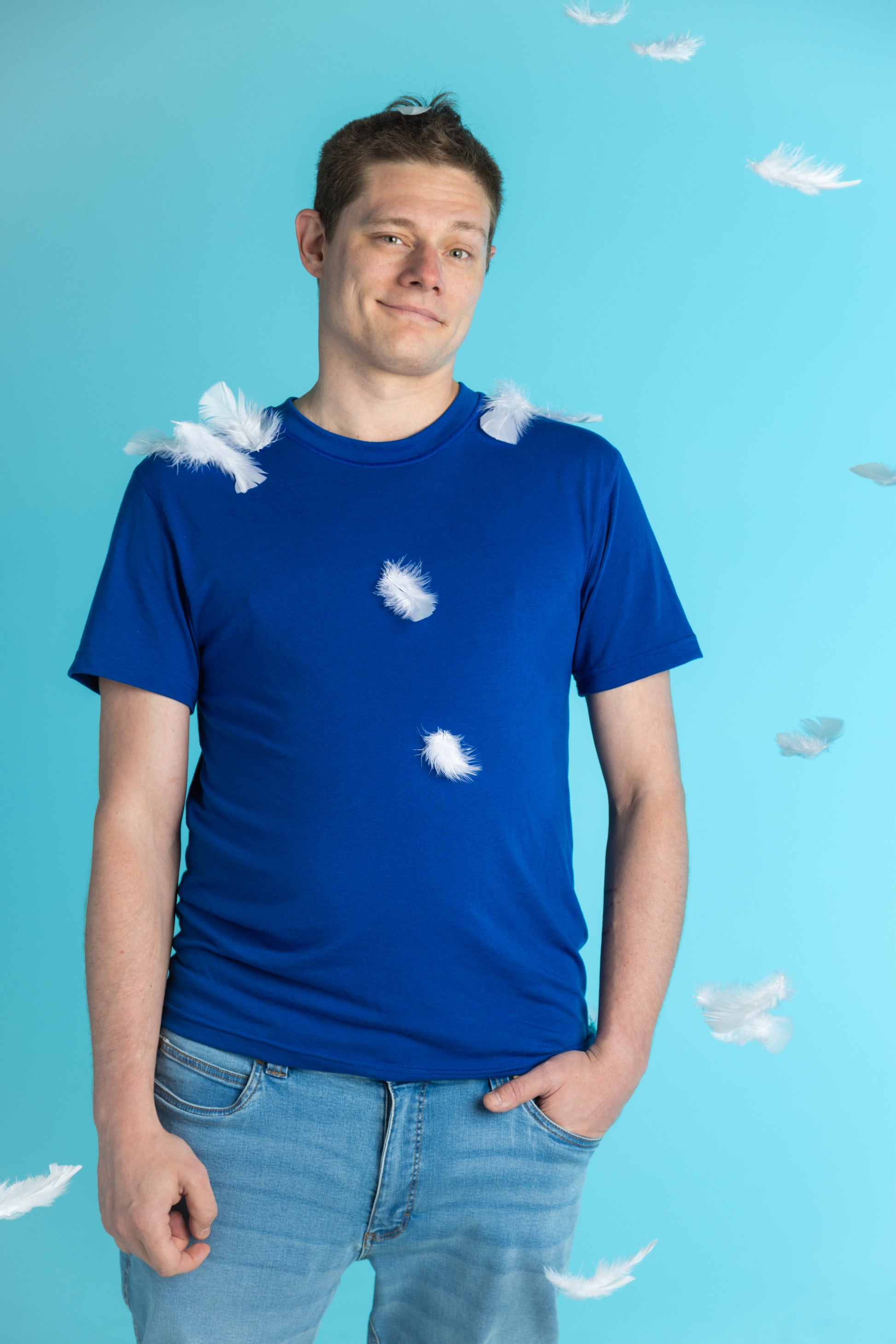 Aqua blue background. A white man with short brown hair wears a navy t-shirt. There are white feathers around him.