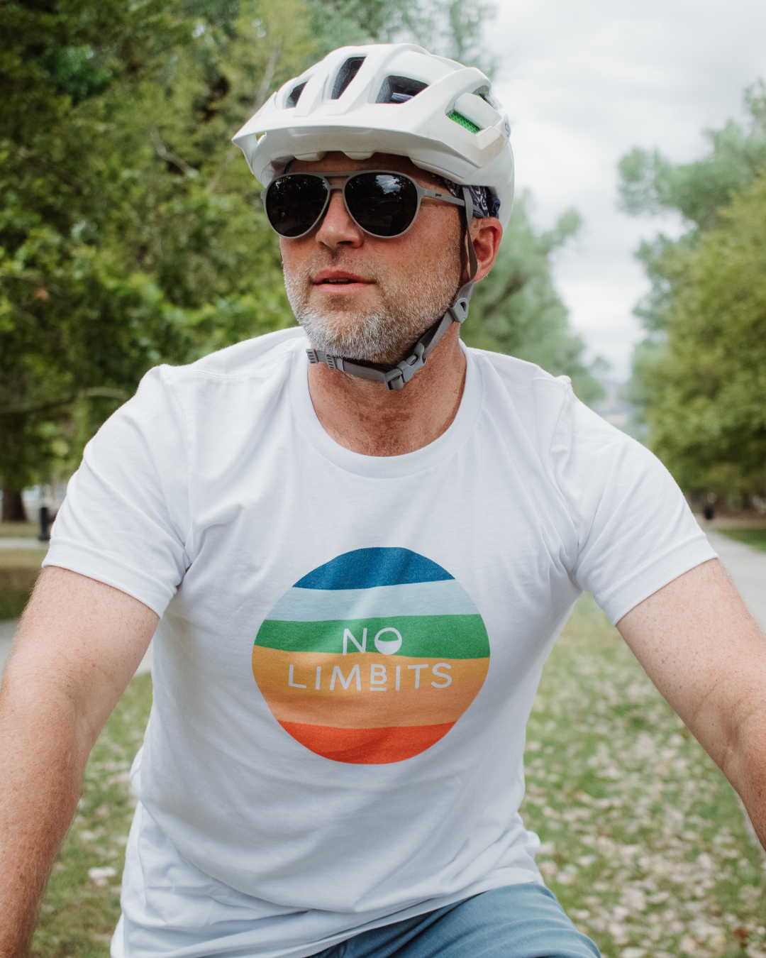 A man riding a bike wearing a white t-shirt that has a rainbow circle on it that says 'No Limbits' The man is wearing a helmet and sunglasses.