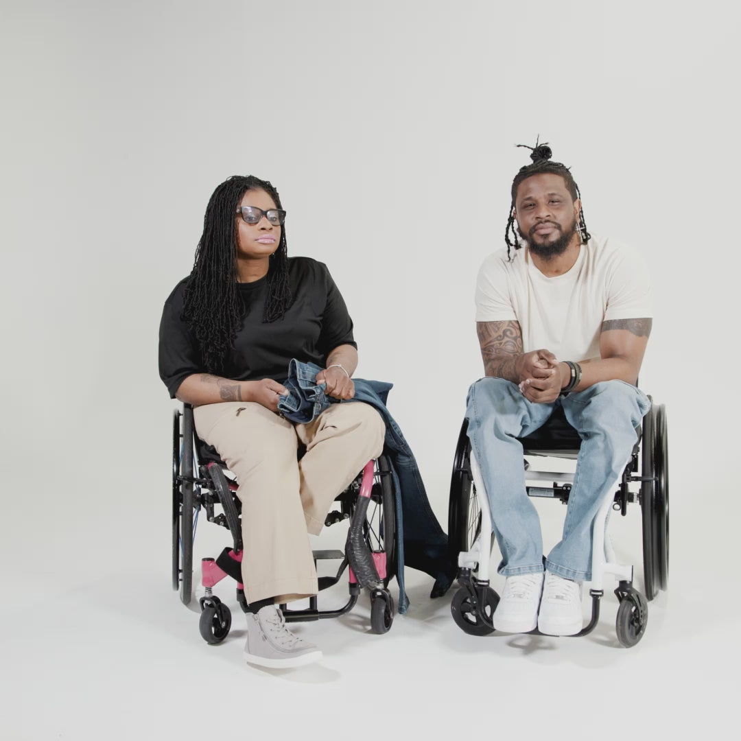 Princess, a Black woman on a pink wheelchair (with long locs, light pink lipstick, clear black sunglasses, and hoops) and Jessi, a Black man on a white wheelchair (with short locs with a bun on the top of his hair, a goatee, cross earrings, and white sneakers) hang out together and demonstrate the wheelchair pants. 