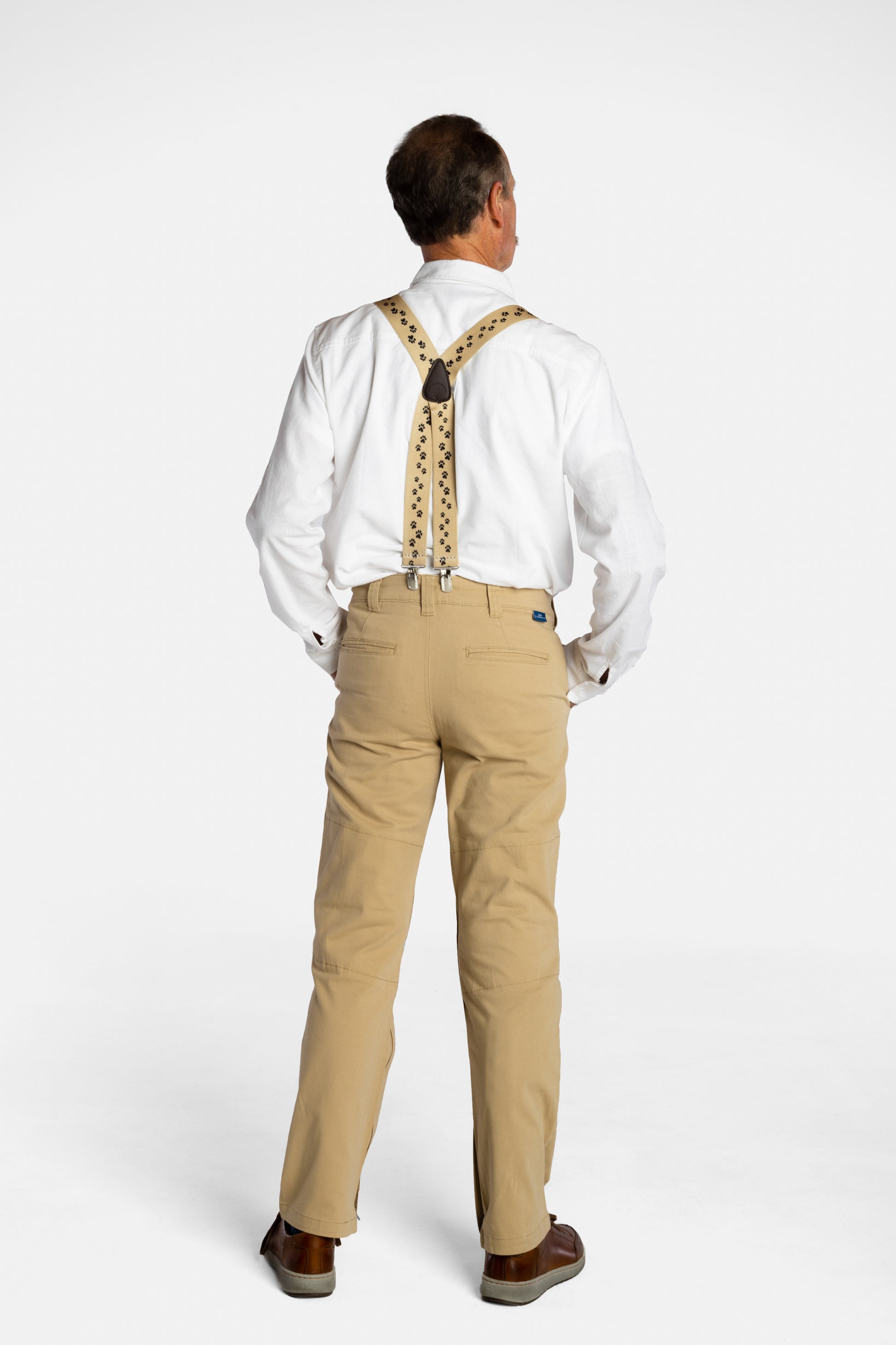 The back of a white, elderly man with a mustache wearing the No Limbits Adaptive Men's Khaki Unlimbited Pants, with a white shirt, tan suspenders, and brown shoes. 