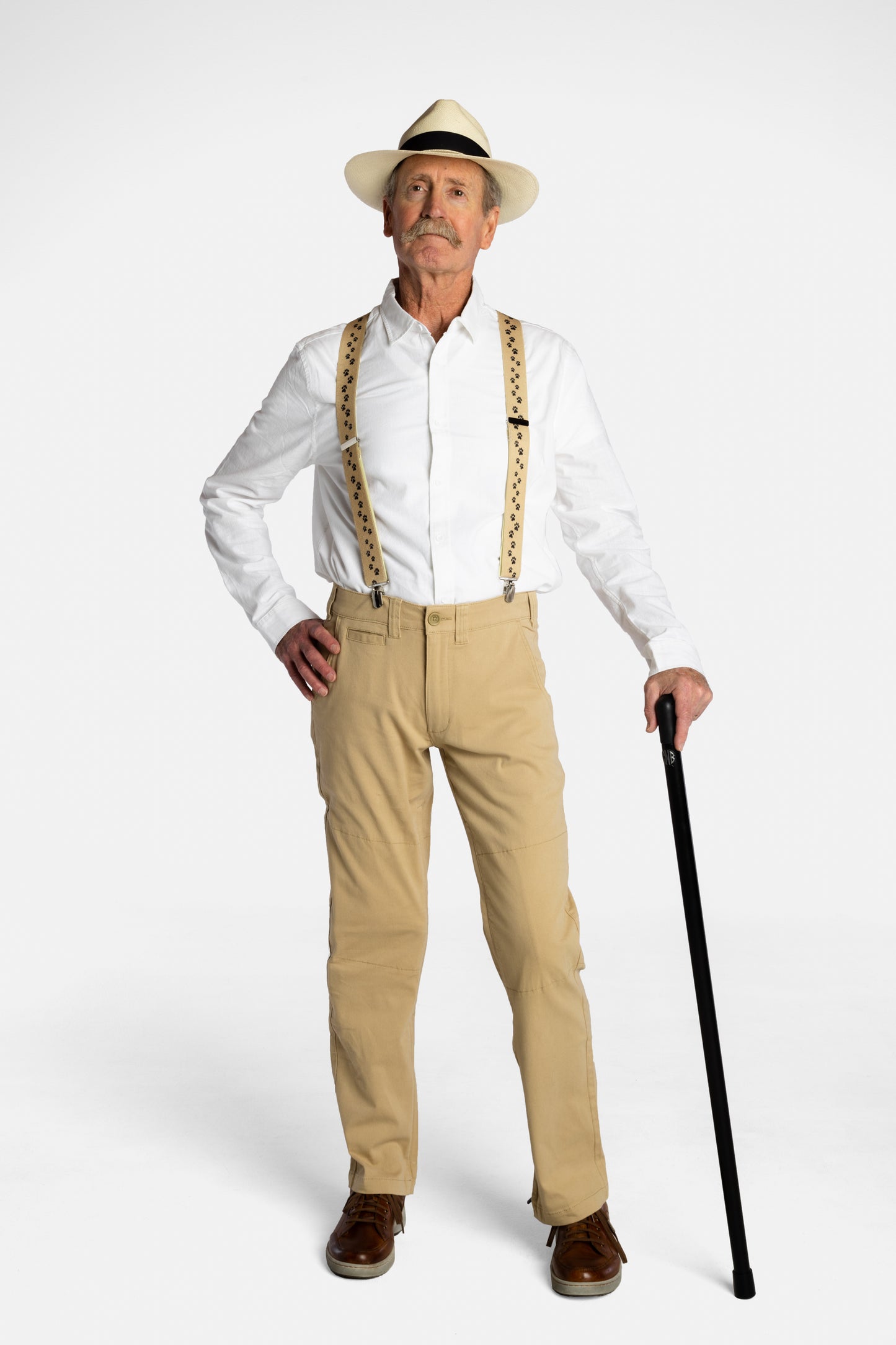 A white, elderly man with a mustache wears the No Limbits Adaptive Men's Khaki Unlimbited Pants with a white shirt, tan suspenders, a matching hat, and brown shoes. He poses with his black cane.