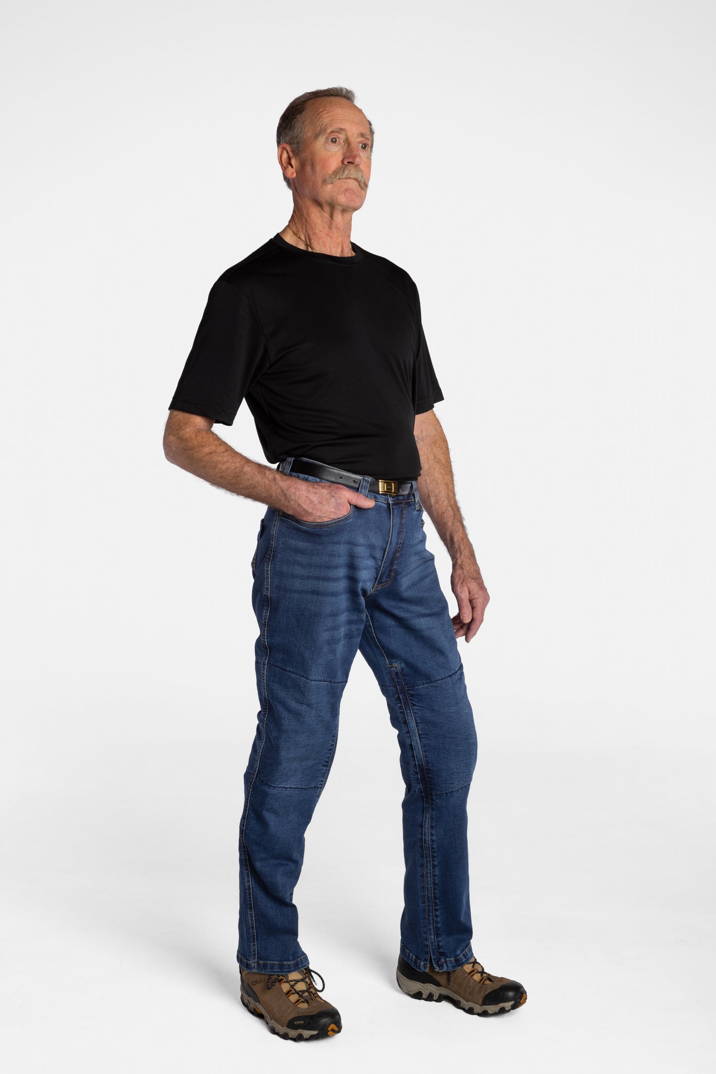 A white, elderly man with a mustache wears the No Limbits Adaptive Men's Dark Wash Unlimbited Pants (which looks like denim jeans), with a black shirt, a brown belt, and brown shoes.