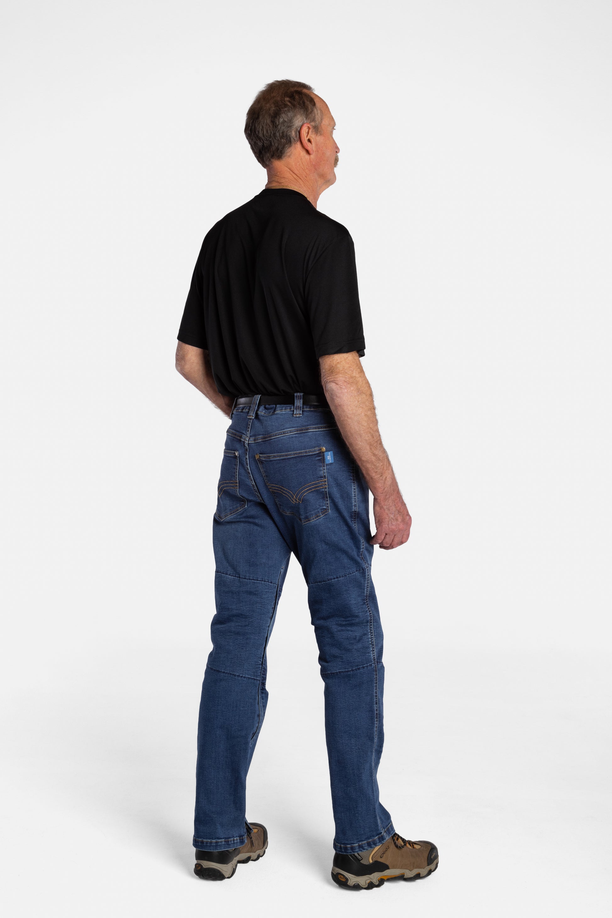 The back of a white, elderly man with a mustache wearing the No Limbits Adaptive Men's Dark Wash Unlimbited Pants (which looks like denim jeans), with a black shirt, a brown belt, and brown shoes.