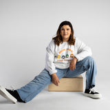 Erica Cole (A white woman with long brown hair) poses in front of a white wall. She sits on a box with a leg stretched forward. She is wearing a white sweater that says “not your inspo with the four No Limbits crew members (Colorful circles with forearm crutches, a prosthetic leg, and a wheelchair), denim jeans, and black & white sneakers.