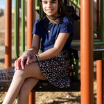 A little girl is wearing a navy blue crewneck t-shirt. She is wearing a black and white skirt with black and white shoes. She is also sitting on a jungle gym. The colors of the jungle gym are orange, green and black.