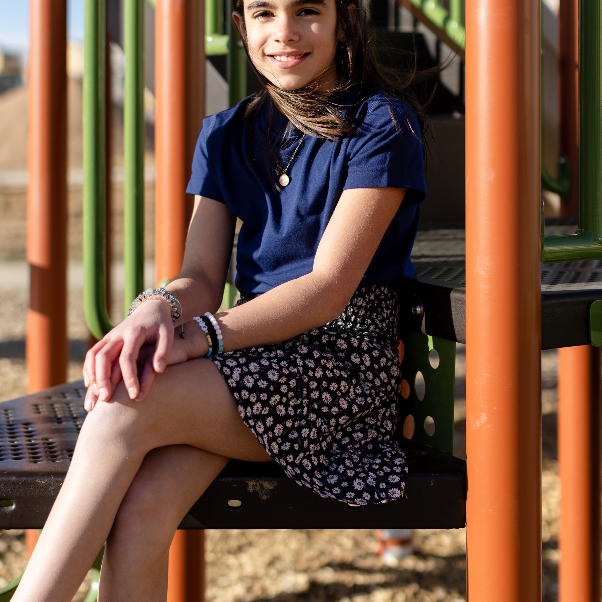 A little girl is wearing a navy blue crewneck t-shirt. She is wearing a black and white skirt with black and white shoes. She is also sitting on a jungle gym. The colors of the jungle gym are orange, green and black.