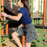 A little girl is wearing a navy blue crewneck t-shirt. She is wearing a black and white skirt with black and white shoes. She is also climbing on a jungle gym. The colors of the jungle gym are orange, green and black.