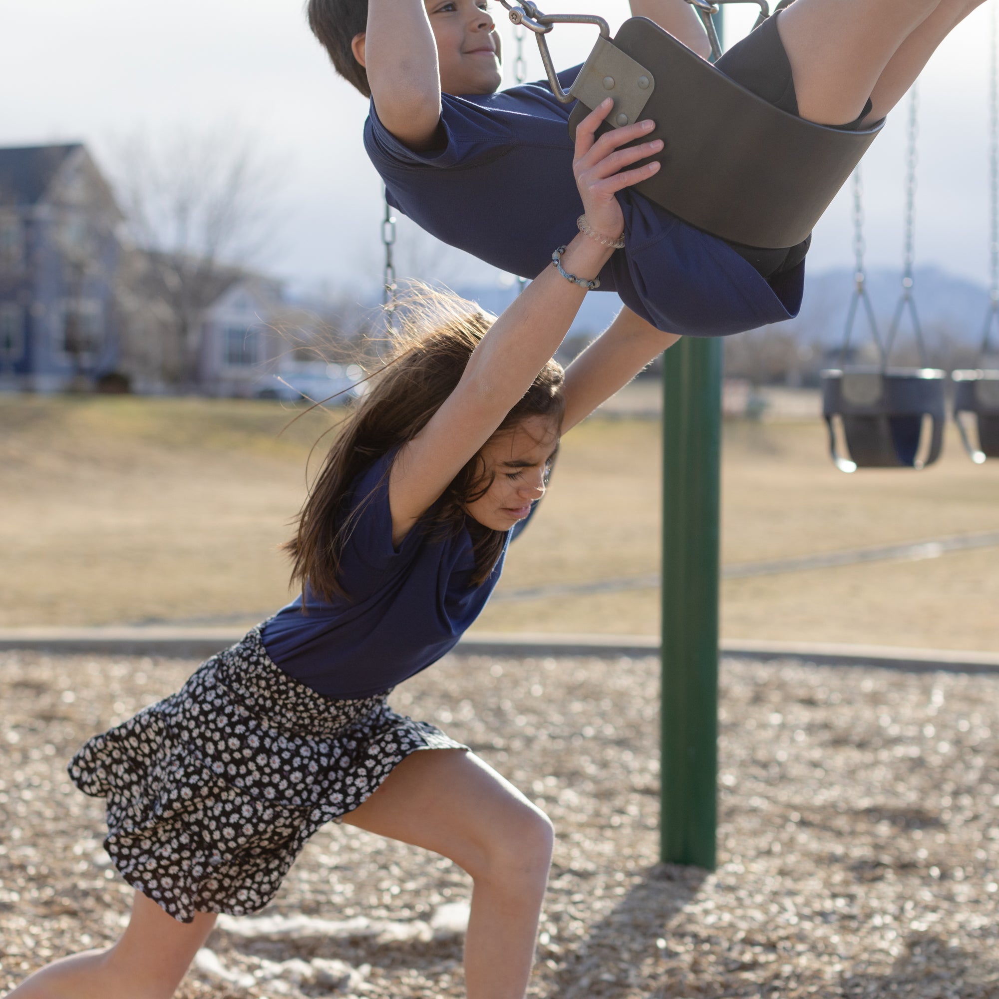 Two kids are playing on the swings together. The little girl is pushing the little boy who is sitting on the black swings. They are both wearing a navy blue basic t-shirt. The little girl is wearing a black and white skirt, and the boy is wearing black shorts.